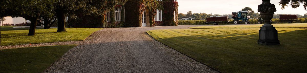 Blick auf das Chateau mit Bäumen am Bildrand und dem Erntevorgang im Hintergrund.
