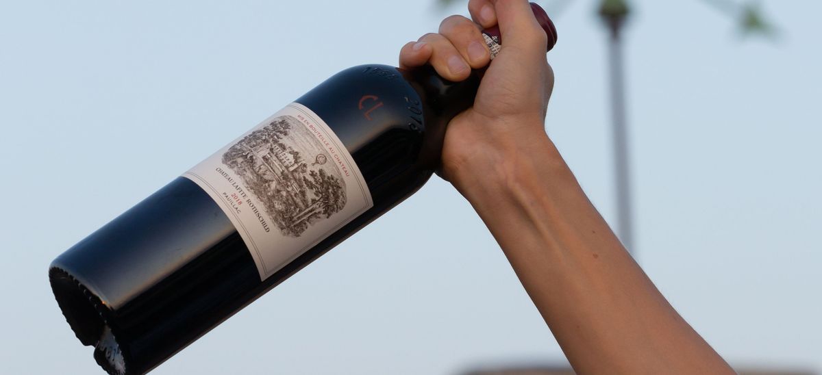 Eine Rotweinflasche von Château Lafite Rothschild wird von einer Hand in den blauen Himmel bei Abendstimmung gehalten.