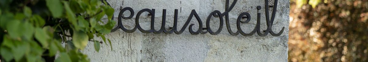 Schriftzug Beausoleil aus Metall an einer Steinsäule und am linken Rand Blätter und im Hintergrund Bäume.