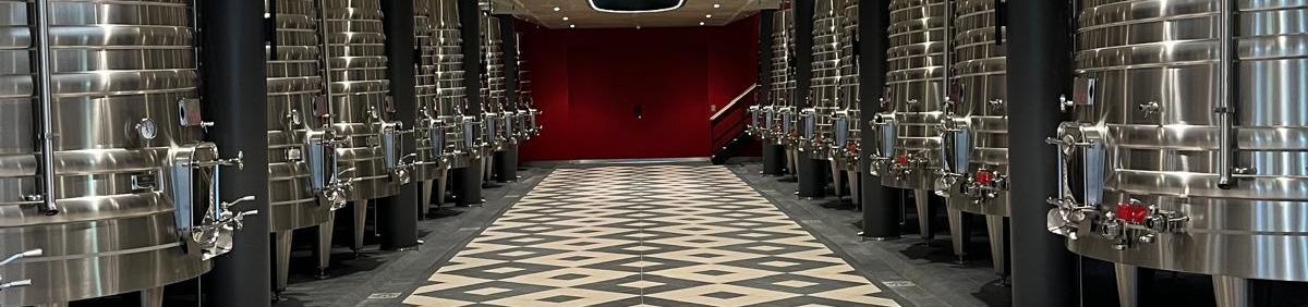 Weinkeller von Château Grand Puy-Lacoste mit Edelstahltanks rechts und links und einer roten mittig platzierten Tür und einem Fliessenboden in Schachbrettoptik