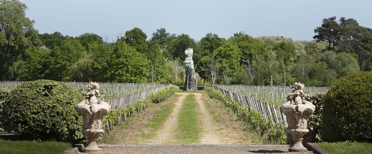 Kiesweg zwischen Weinreben mit Statue am Ende des Weges und Buschen am Anfang