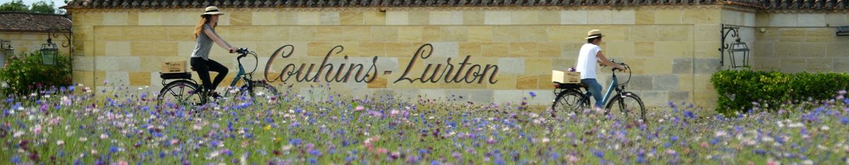 Zwei Personen fahren entlang des Anwesens von Château Couhins-Lurton Fahrrad, davor sieht man eine Blumenwiese.