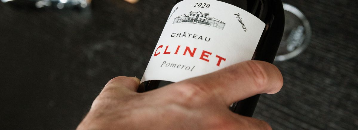 Eine Hand hält eine Flasche Château Clinet 2020 un schenkt Rotwein in ein Weinglas ein.