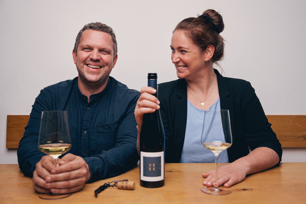 Mann und Frau sitzen lachend hinter Holztisch mit Weinflasche von Weingut Hees in der Hand