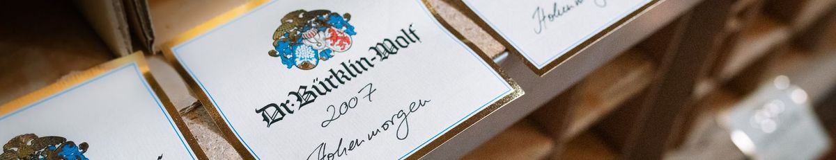 Weinflaschen des Weinguts Dr. Bürklin-Wolf liegen in einem Weinregal, vor jeder Flasche ist das passende Etikett zum Wein abgelegt