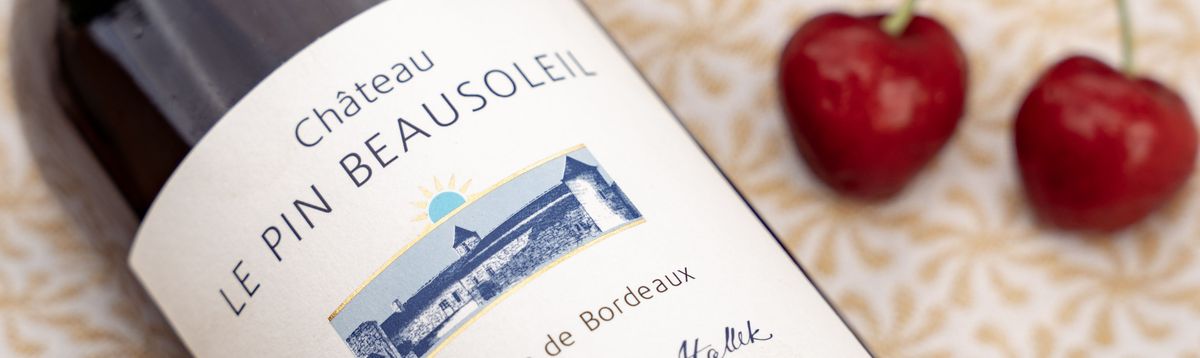 Eine Rotweinflasche von Château Le Pin Beausoleil 2019 auf einer Tischdecke mit zwei Kirschen daneben.