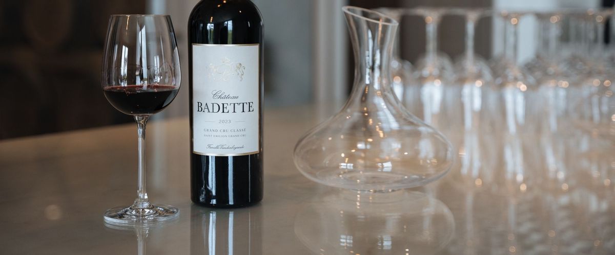 Rotweinflasche von Chateau Badette und eingeschenktes Glas auf einem Tisch zusammen mit weiteren leeren Weingläsern und einem leeren Decanter.